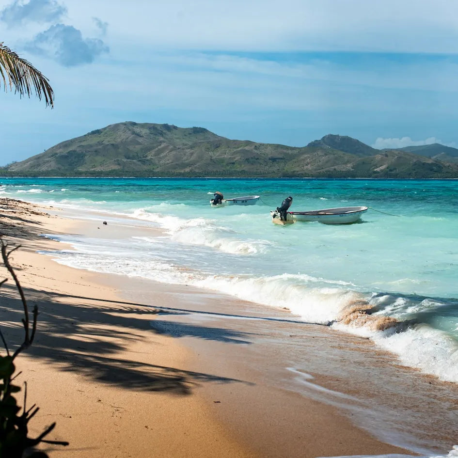 Beach scene with clear blue water, white waves, and a small Fiji island in the distance.