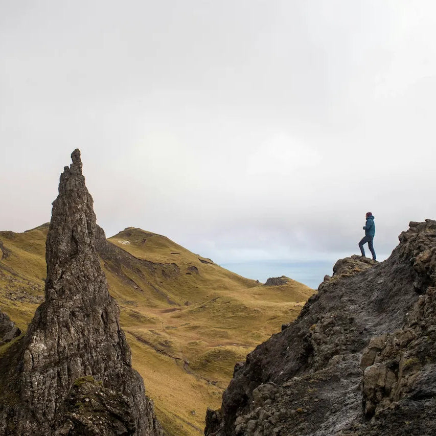 Person standing on a rocky outcrop in a mountainous landscape with a cloudy sky.